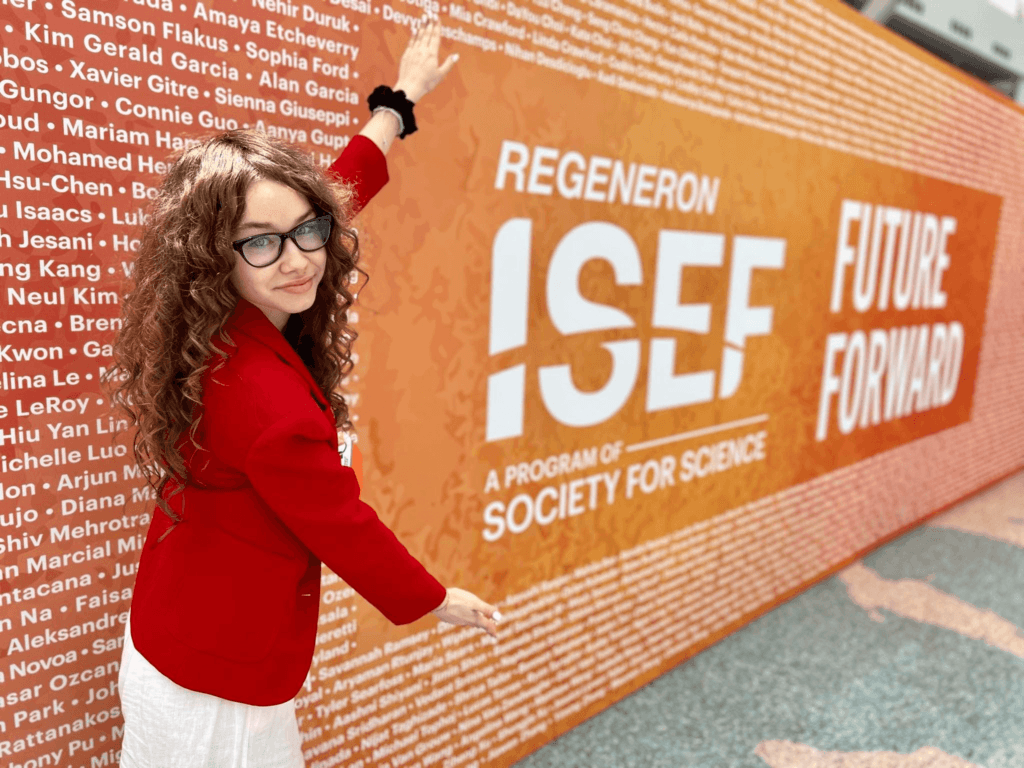 Kornelia Wieczorek, młoda innowatorka z Polski, pozuje na tle ściany z nazwiskami uczestników Regeneron ISEF 2024. Ubrana w czerwony żakiet, wskazuje na swoje nazwisko na tablicy.
