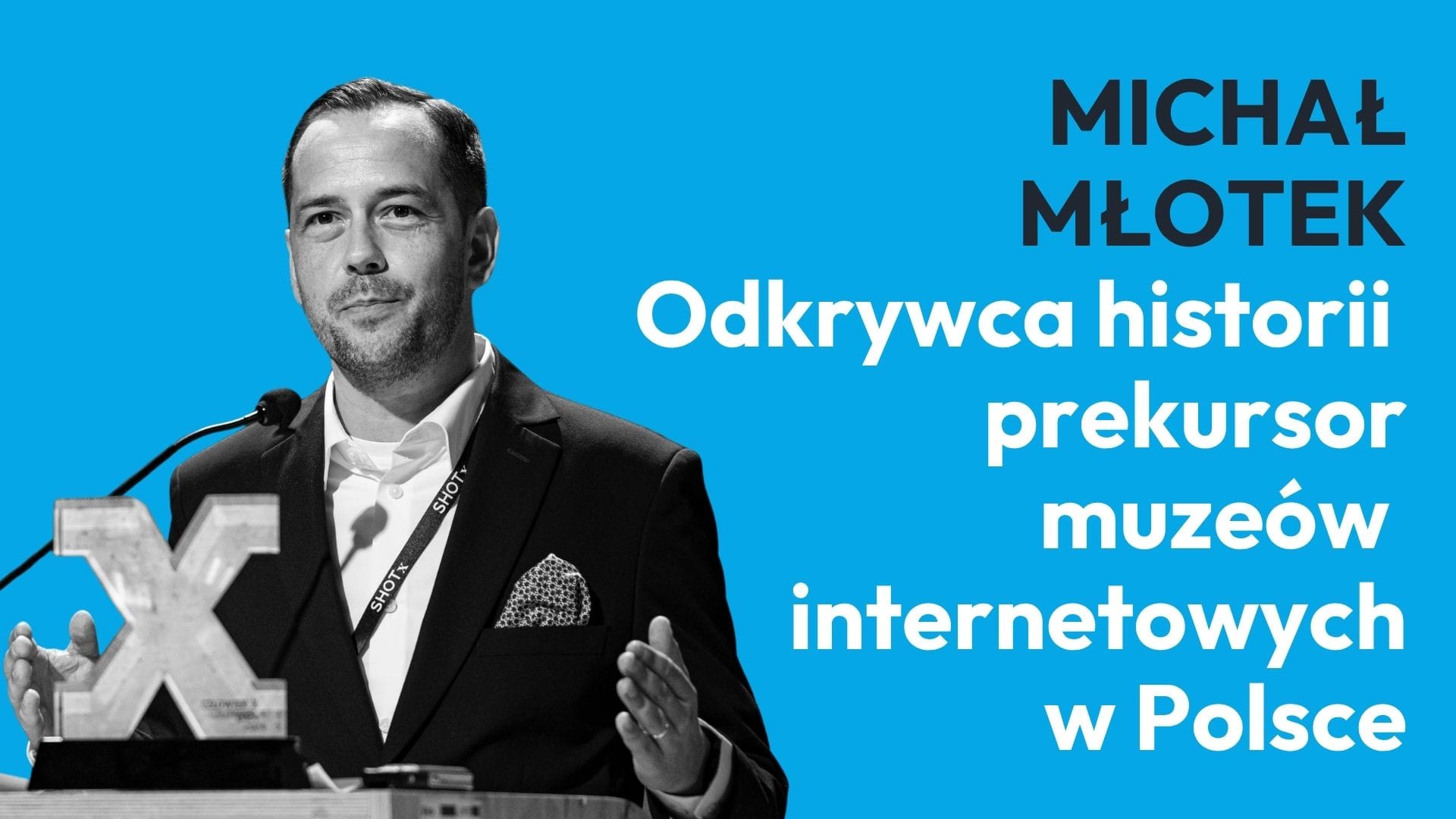 Mężczyzna w garniturze mówi do mikrofonu przy podium na tle jasnoniebieskiego plakatu z dużymi białymi napisami po prawej stronie.