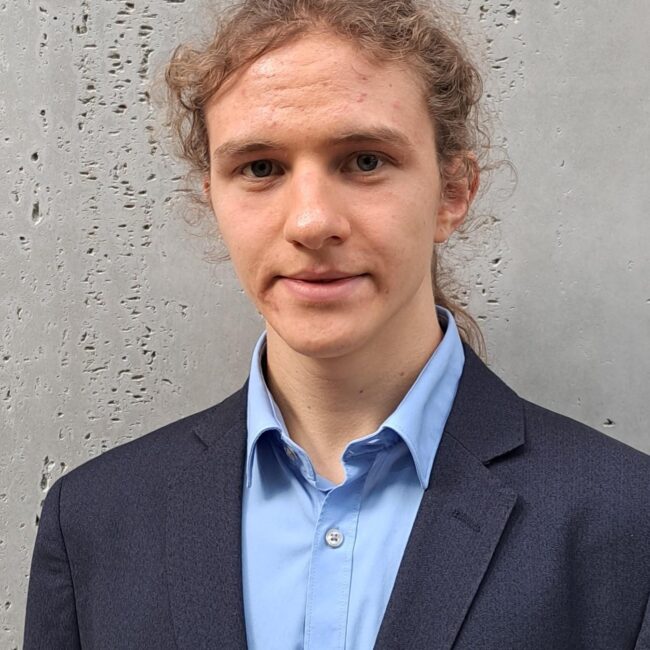 Portrait of a young man in a navy blazer and light blue shirt standing against a gray textured wall.